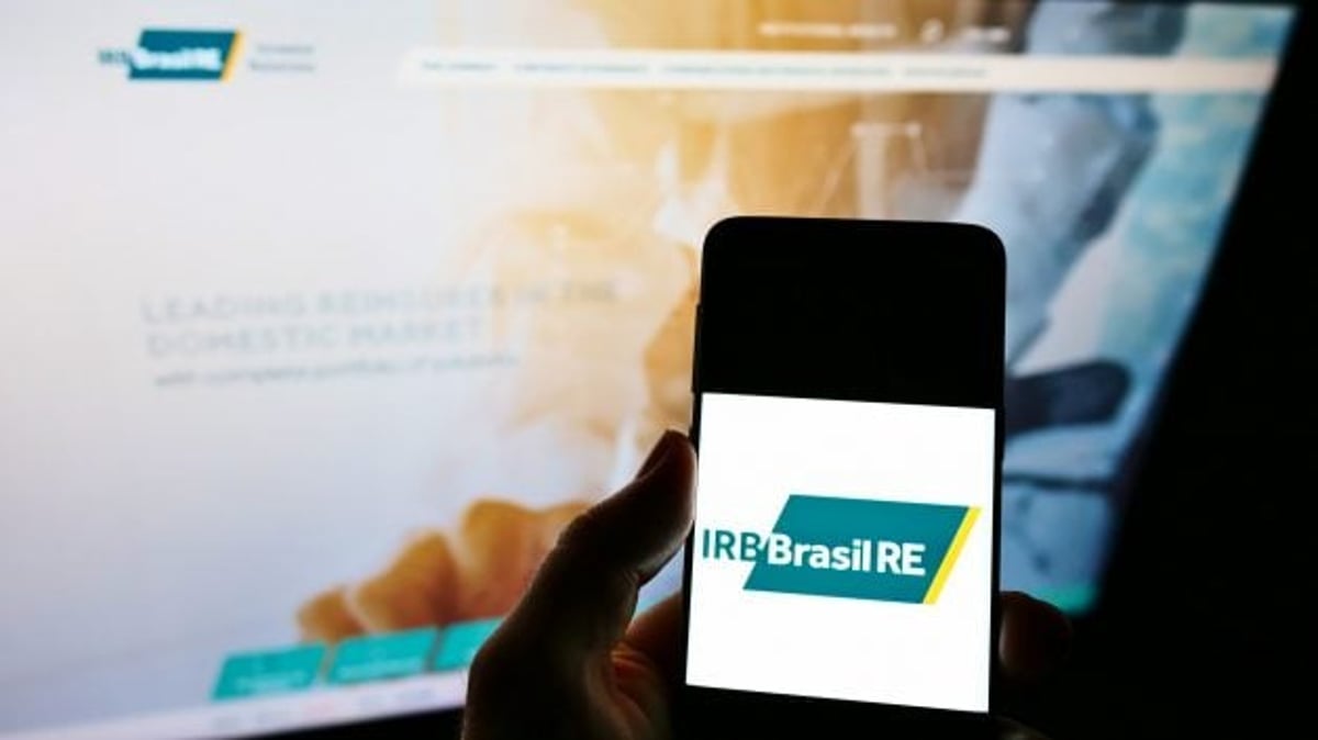 Imagem de um celular com o logo do IRB (IRBR3) sendo exibido na tela