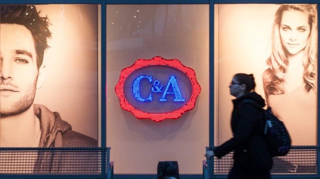 Fachada de loja da C&A (CEAB3). Uma mulher vestida de preto caminha no primeiro plano; o logo da empresa, nas cores vermelho, azul e branco, aparece ao fundo