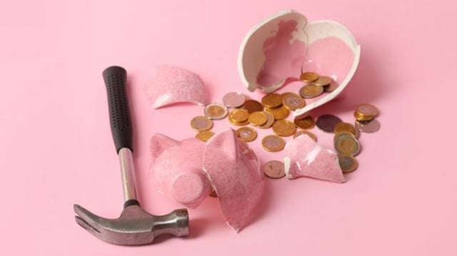 Cofrinho rosa em formato de porco quebrado, com moedas e um martelo ao seu redor, representando poupança