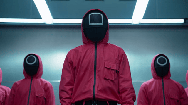 Imagem da série coreana Round 6, sucesso global da Netflix (NFLX34). Nela, três pessoas encapuzadas e vestidas de vermelho, com máscaras com símbolos geométricos, aparecem paradas em frente a uma parede