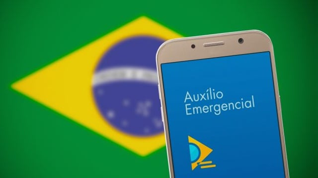 Imagem de um celular com o aplicativo do auxílio emergencial. Em segundo plano, uma bandeira do Brasil desfocada