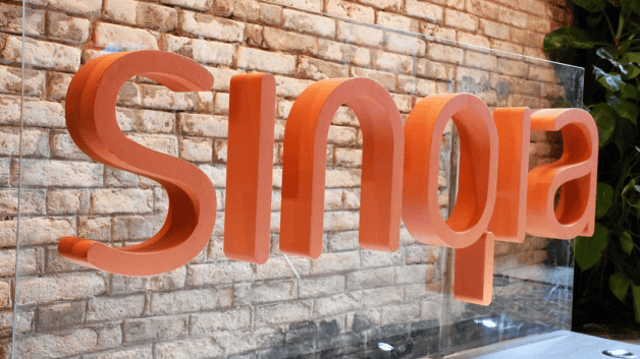 Imagem com a palavra 'Sinqia' (SQIA3) escrita em letras laranjas. Ao fundo, uma parede de tijolos