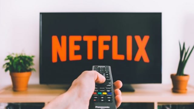Imagem mostrando uma mão segurando um controle remoto apontado para uma TV; na tela, aparece o logo da Netflix (NFLX34)