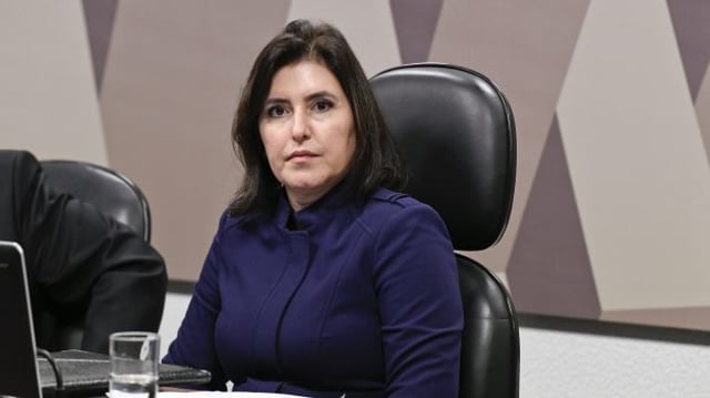 Simone Tebet veste blusa azul marinho. Ela está sentada em uma cadeira preta, com um notebook em sua frente e um copo de água ao lado.