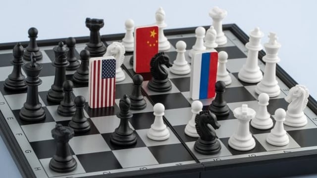 Tabuleiro de xadrez; nele, há três peças diferentes, identificadas com as bandeiras dos EUA, da China e da Rússia; simboliza a tensão geopolítica e a guerra no leste europeu