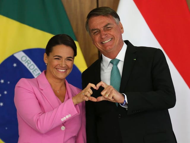 A presidente da Hungria veste rosa e faz um coração com uma das mãos junto com o presidente Jair Bolsonaro. Bandeiras de Hungria e Brasil ao fundo