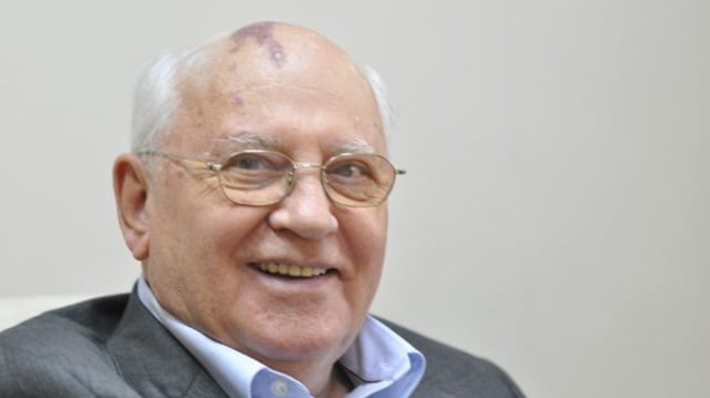 Foto de meio corpo de Mikhail Gorbachev, usando óculos e sorrindo, vestindo terno cinza e camisa azul