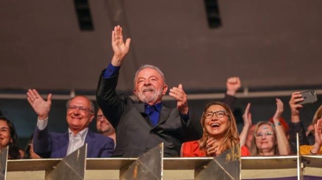 Lula em pé, em palanque, batendo palmas. Ao lado dele, Geraldo Alckmin dá tchau e do outro lado, a esposa de Lula, Janja, aplaude