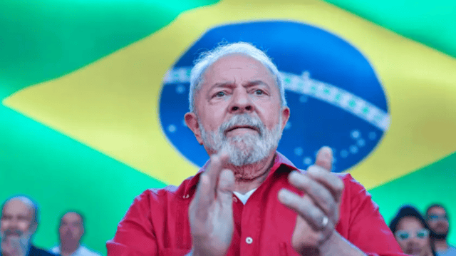 Lula veste camisa vermelha, bate palmas, em um cenário com bandeira do Brasil ao fundo