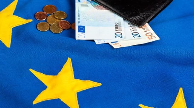 Bandeira da zona do euro com cédulas e moedas de euro no topo; a Europa é um dos destaques da agenda econômica da semana