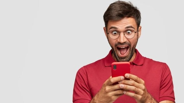 Photo of amazed European blogger holds modern cell phone, recieves unexpected message from followers, connected to wireless internet, isolated over white background. Reaction and technology concept