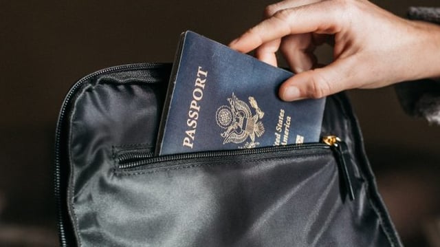 Mão segurando um passaporte azul, coloca o passaporte dentro de uma bolsa preta de viagem