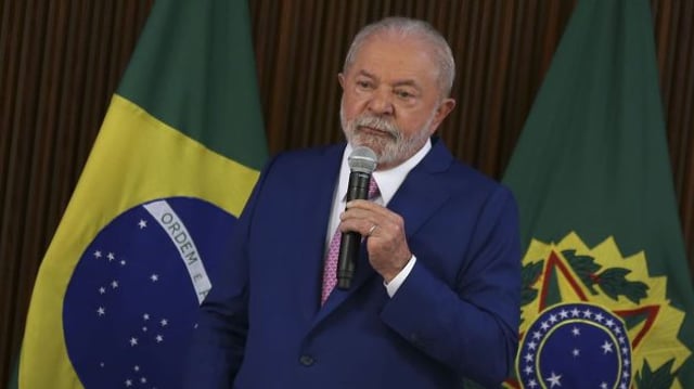 Homem de terno azul marinho, fala de pé, com microfone a mão. Bandeira do Brasil ao fundo do cenário.