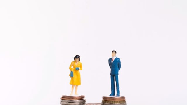 homem com terno azul e mulher de vestido amarelo, ambos em pé, cada um sobre uma torre de moedas representando a igualdade salarial