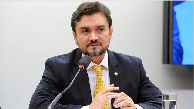 Foto de meio corpo de homem vestindo terno escuro, camisa branca e gravata amarela. Um microfone está na frente ele, que tem barba.