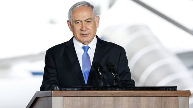 O primeiro-ministro de Israel, Benjamin Netanyahu, veste terno azul e gravata azul clara e está de pé de frente a um microfone