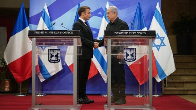 O presidente da França, Emmanuel Macron, aperta as mãos do primeiro-ministro de Israel, Benjamin Netanyahu. Ambos estão de pé, com bandeiras de seus países ao fundo.