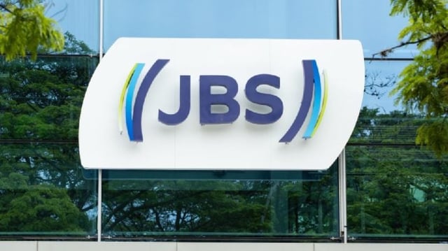 Logo da companhia JBS, ao fundo céu azul e árvores espelhados na fachada do prédio