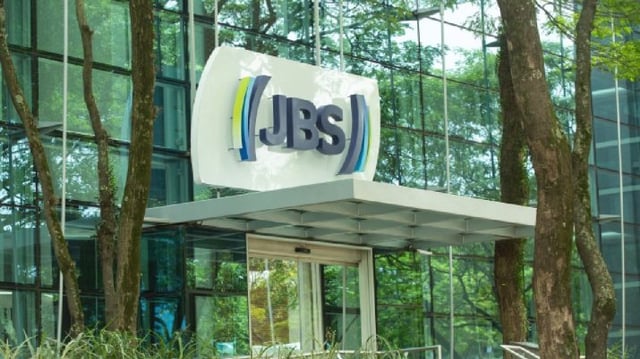 Logo da companhia JBS, ao fundo céu azul e árvores espelhados na fachada do prédio