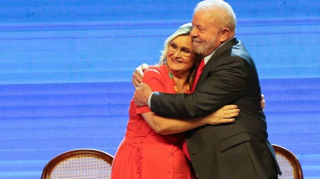 Rita Serrano, então presidente da Caixa, abraça o presidente Lula. Ela veste um vestido vermelho e ele, terno escuro.