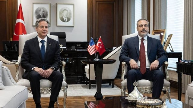 O secretário de Estado dos EUA, Antony J. Blinken, está sentado em uma cadeira ao lado do ministro das Relações Exteriores da Turquia, Hakan Fidan. Entre os dois, uma mesa com as bandeiras dos EUA e da Turquia.