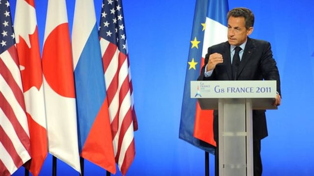 O ex-presidente da França, Nicolas Sarkozy, discursa em frente a um púlpito, com bandeiras de várias países atrás. O fundo da imagem é azul.