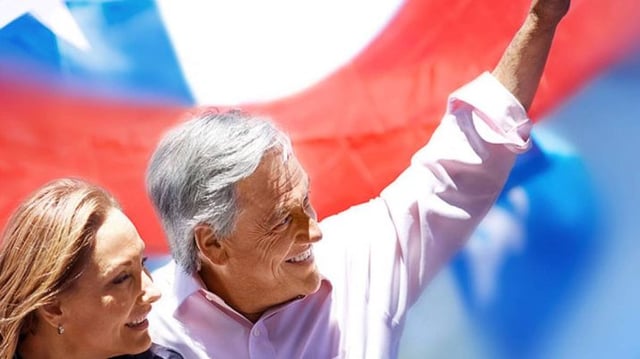 Homem abraçado com uma mulher, com a bandeira do Chile ao fundo, acena com um dos braços levantados