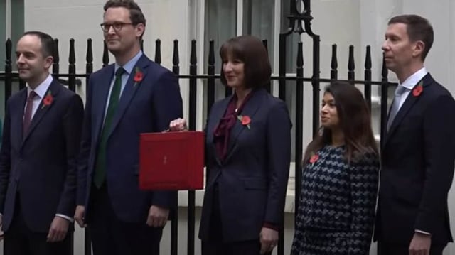 A ministra das Finanças do Reino Unido, Rachel Reeves, aparece de pé em frente a um portão, com uma mala vermelha nas mãos. Ela veste terno azul marinho e está ladeada de membros da sua equipe de governo