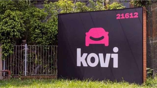 Placa com fundo preto e a marca da Kovi, empresa de app de transporte, em branco e o desenho de um carro em vermelho. A placa está instalada em um gramado.