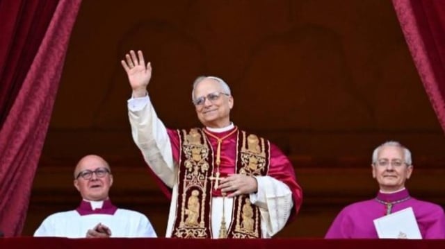 Imagem do novo papa Leão 14, que está ao centro, com as vestes papais em branco, vermelho e dourado. Cardeais vestindo roxo estão ao lado dele.