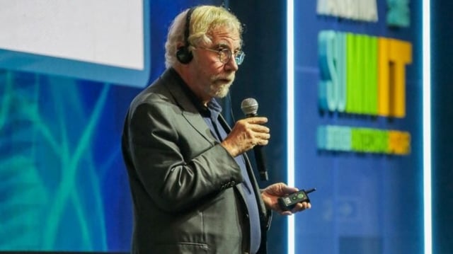 Paul Krugman, idoso branco, falando com microfone em cima do palco