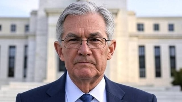 O presidente do Federal Reserve, Jerome Powell, está parado em frente à sede do banco central norte-americano. Ele usa óculos e veste terno azul marinho e camisa branca.