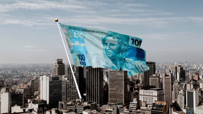 Montagem mostra uma "bandeira" com uma nota de 100 reais no meio de uma cidade cheia de prédios