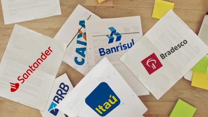 Logos do Bradesco, Itaú Unibanco, Santander, Caixa, Banrisul e BRB em papeis jogados em cima da mesa