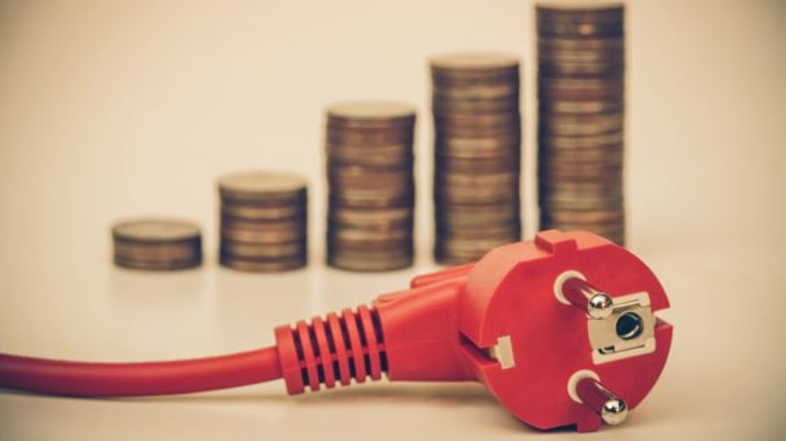 Imagem mostra uma tomada vermelha em primeiro plano com pilhas de moedas ao fundo que representam o preço caro da energia