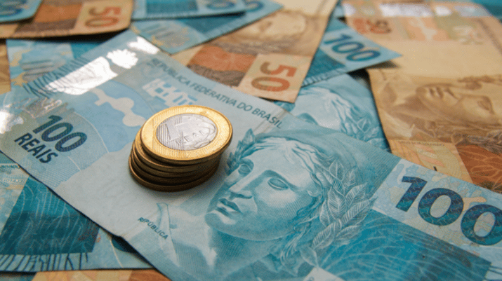 Notas de 100 e 50 reais espalhadas, com uma pilha de moedas de 1 real ao centro