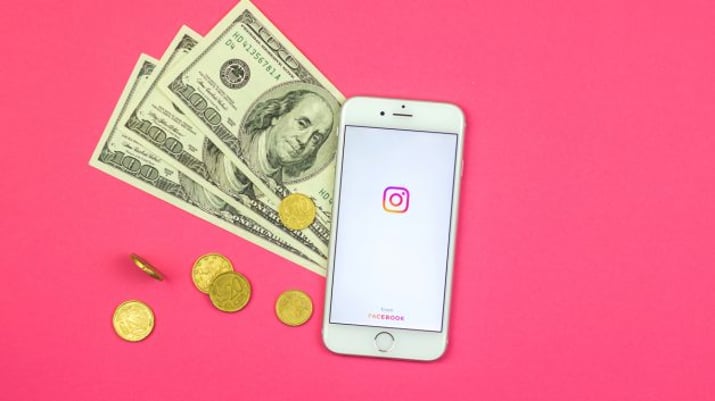 Fundo rosa com um celular aberto no Instagram e cédulas e moedas ao lado