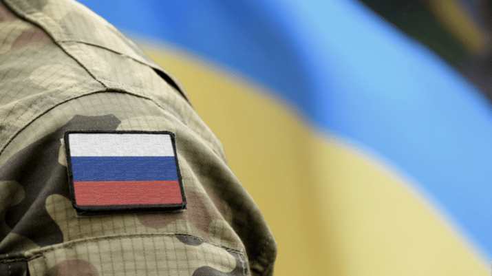 Parte do braço de um soldado russo, com a bandeira do país no ombro e uma bandeira da Ucrânia ao fundo | Bolsa