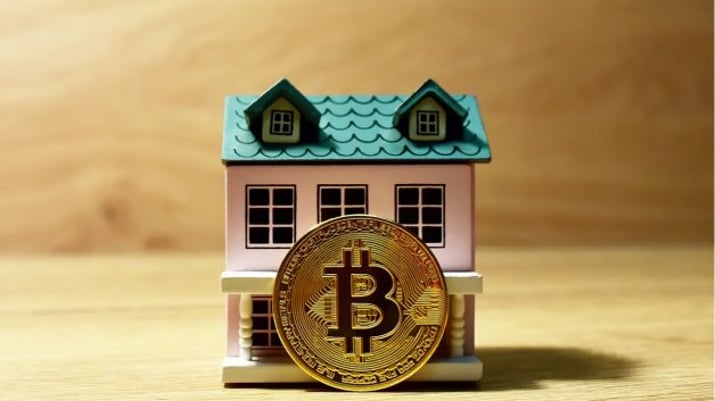 Moeda de bitcoin em frente a uma pequena casa branca com telhado azul