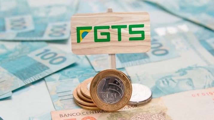 Montagem mostrando cédulas e moedas de real espalhadas numa mesa; no topo, uma placa de madeira com a sigla FGTS, em verde