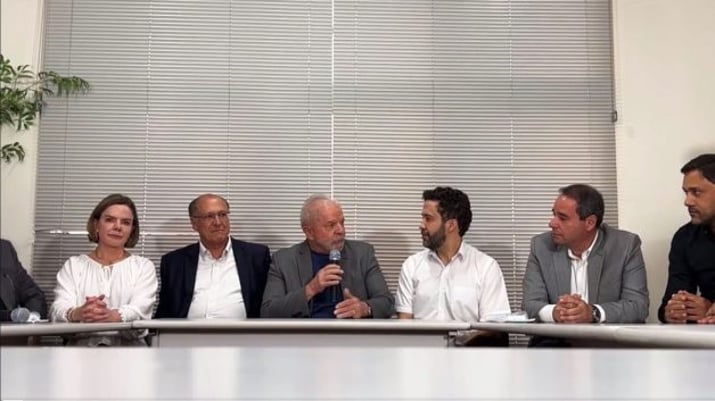 Lula com microfone na mão, sentado em uma mesa ao lado de políticos