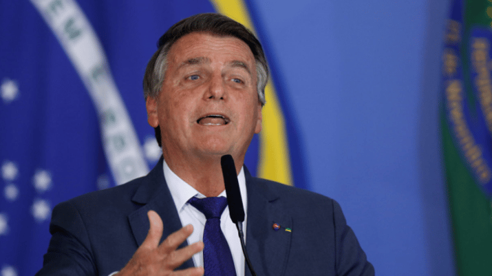 NÃO USAR Bolsonaro veste terno azul e camisa branca. Fala ao microfone durante discurso, com bandeira do Brasil ao fundo.