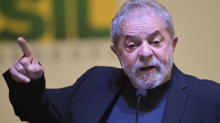 Luiz Inácio Lula da Silva de terno preto e camiseta preta, aponta pra cima enquanto fala ao microfone