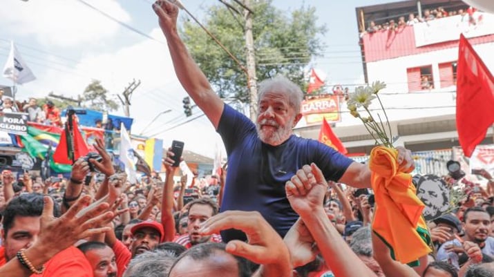 Lula veste camiseta azul marinho, segura uma camiseta laranja e flores brancas em uma mão, a outra está erguida. Ele está nos braços no povo, no meio de uma multidão