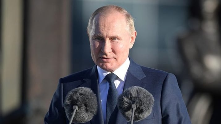 Vladimir Putin vestindo terno azul marinho e camisa branca, em frente a um púlpito com dois microfones na frente