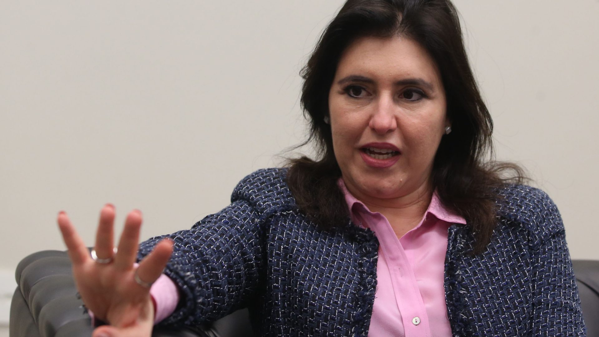 Simone Tebet sentada em sofá, faz o número 4 com as mãos.