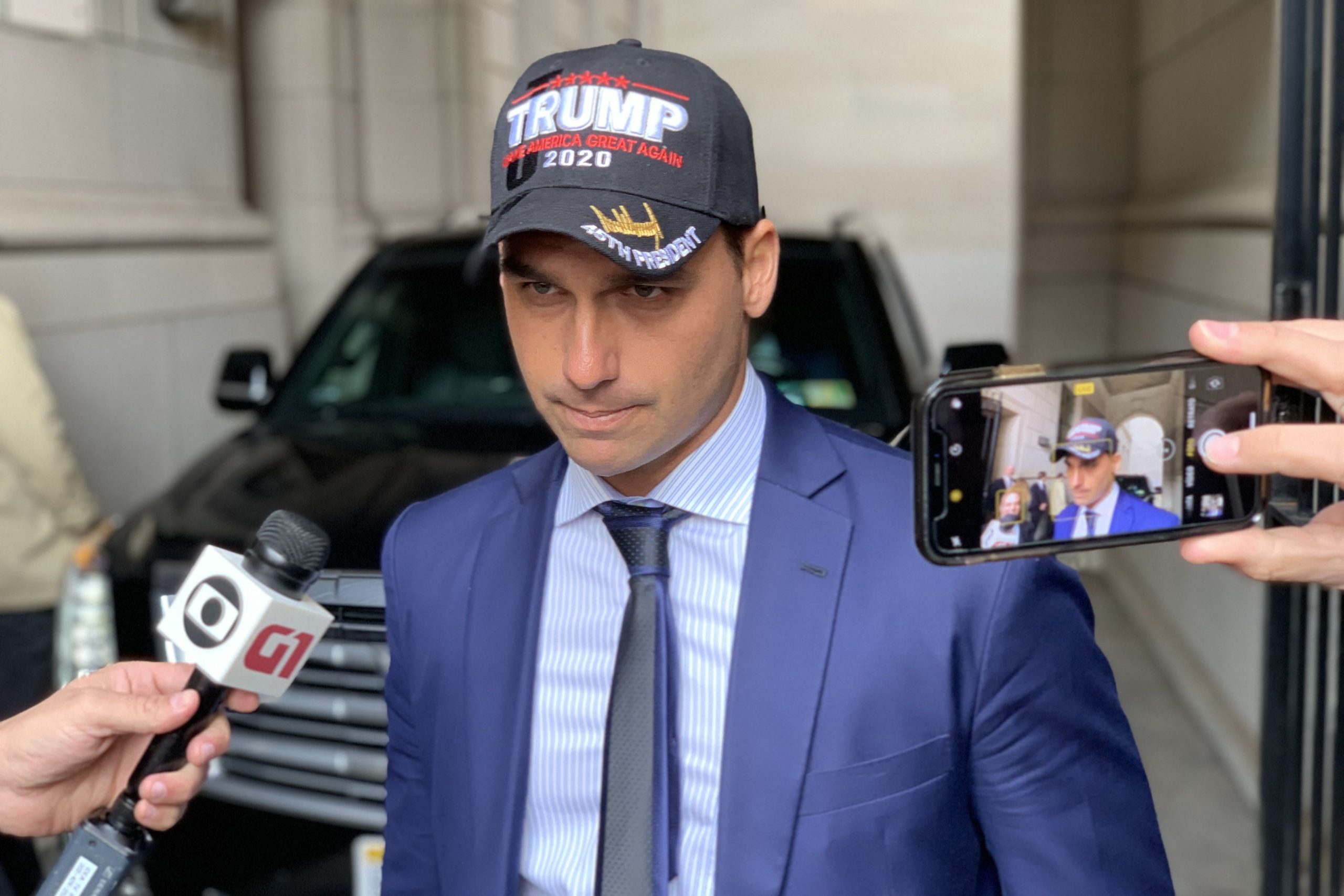 Eduardo Bolsonaro com boné escrito "Trump" no meio de repórteres.