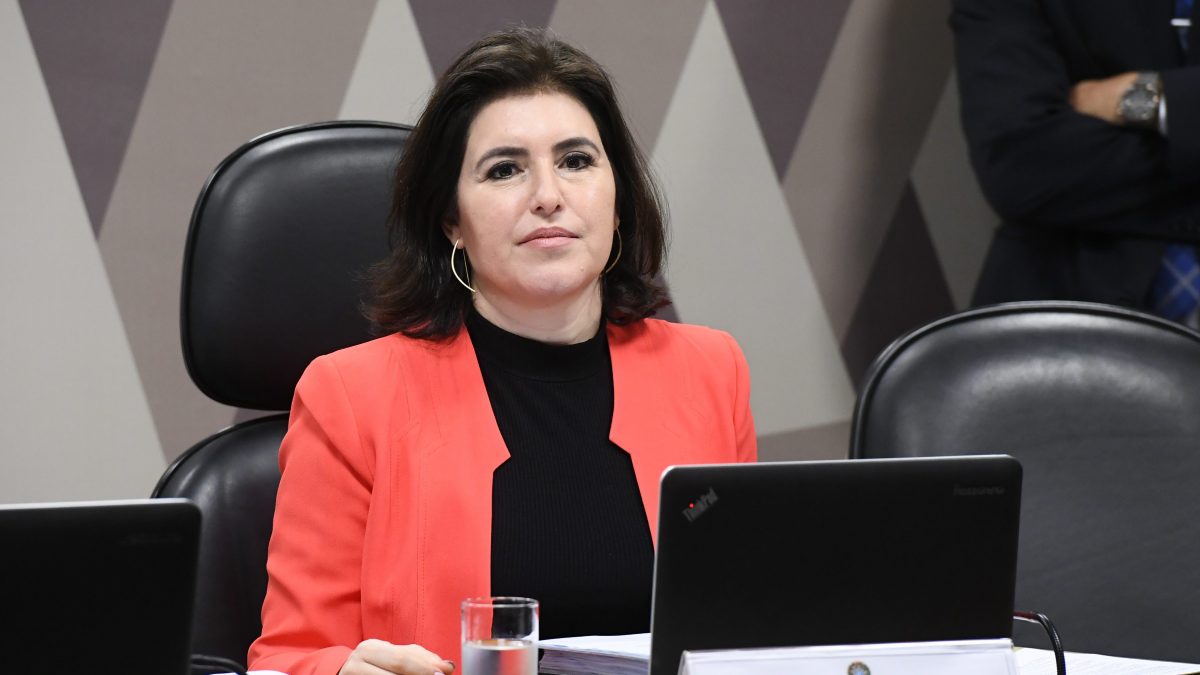 Senadora Simone Tebet está sentada em uma cadeira preta, com notebook na frente. Ela veste blusa preta e casaco laranja.