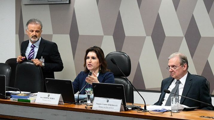 Homem de pé veste terno preto e segura em cadeira preta. No centro, Simone Tebet está sentada com notebook a frente. Do outro lado, homem veste terno cinza escuro. Ele também está sentado