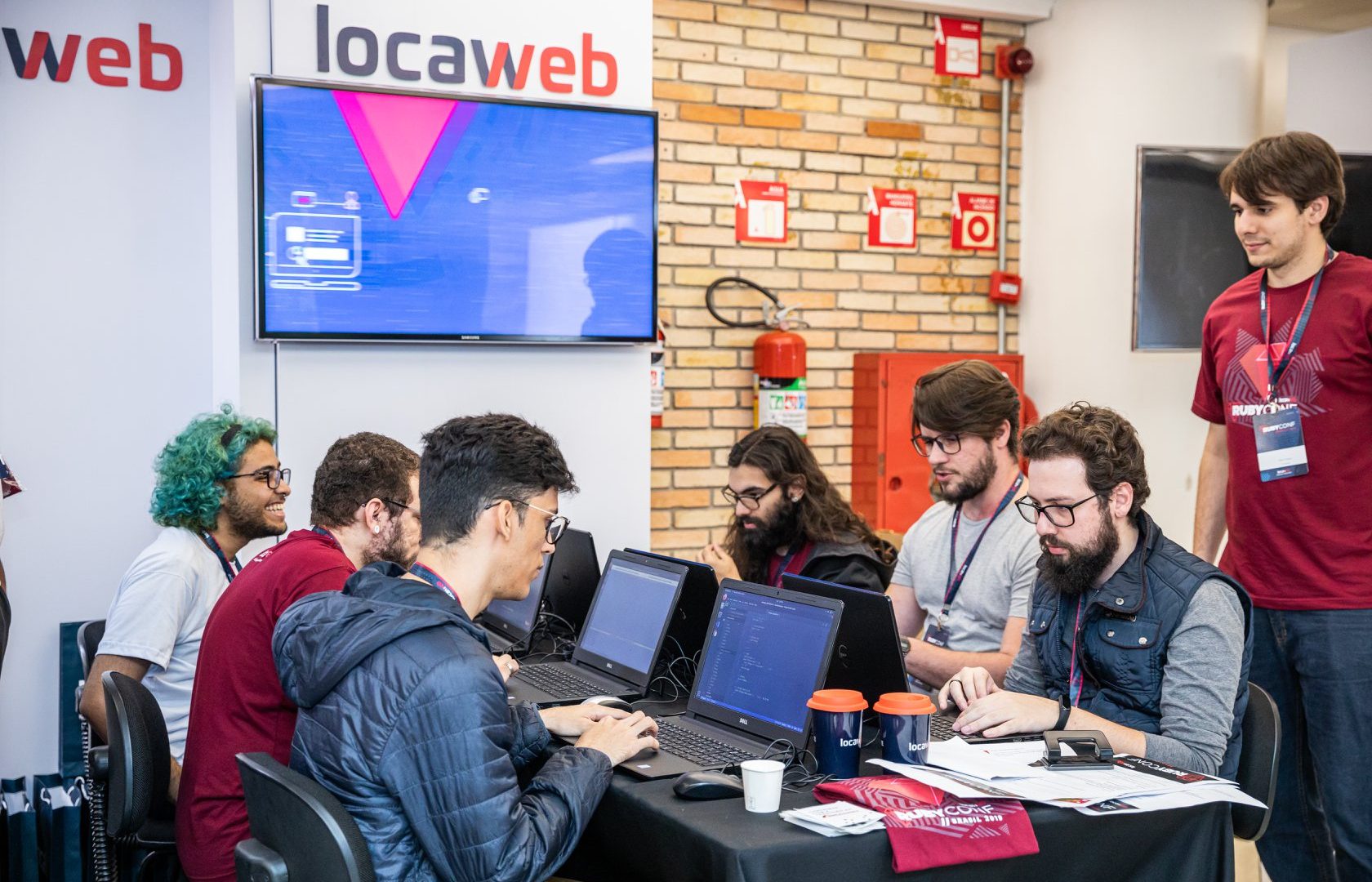Escritório com pessoas em frente a seus computadores. Ao fundo, logo da Locaweb